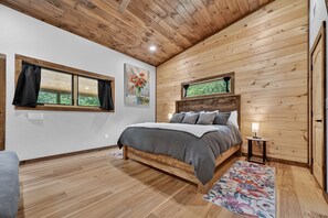 A bedroom with a wooden ceiling, a bed with gray bedding, a nightstand with a lamp, and a window with curtains.