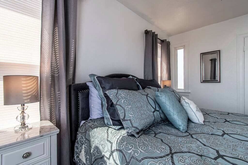 A bedroom with a bed, a bedside table, a lamp, curtains, a window, and a mirror on the wall.