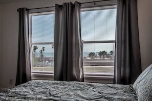A bedroom with a bed, two windows with curtains, and a view of palm trees and a distant landscape. - Tranquil Terrace - Duplex #2 (Corpus Christi)