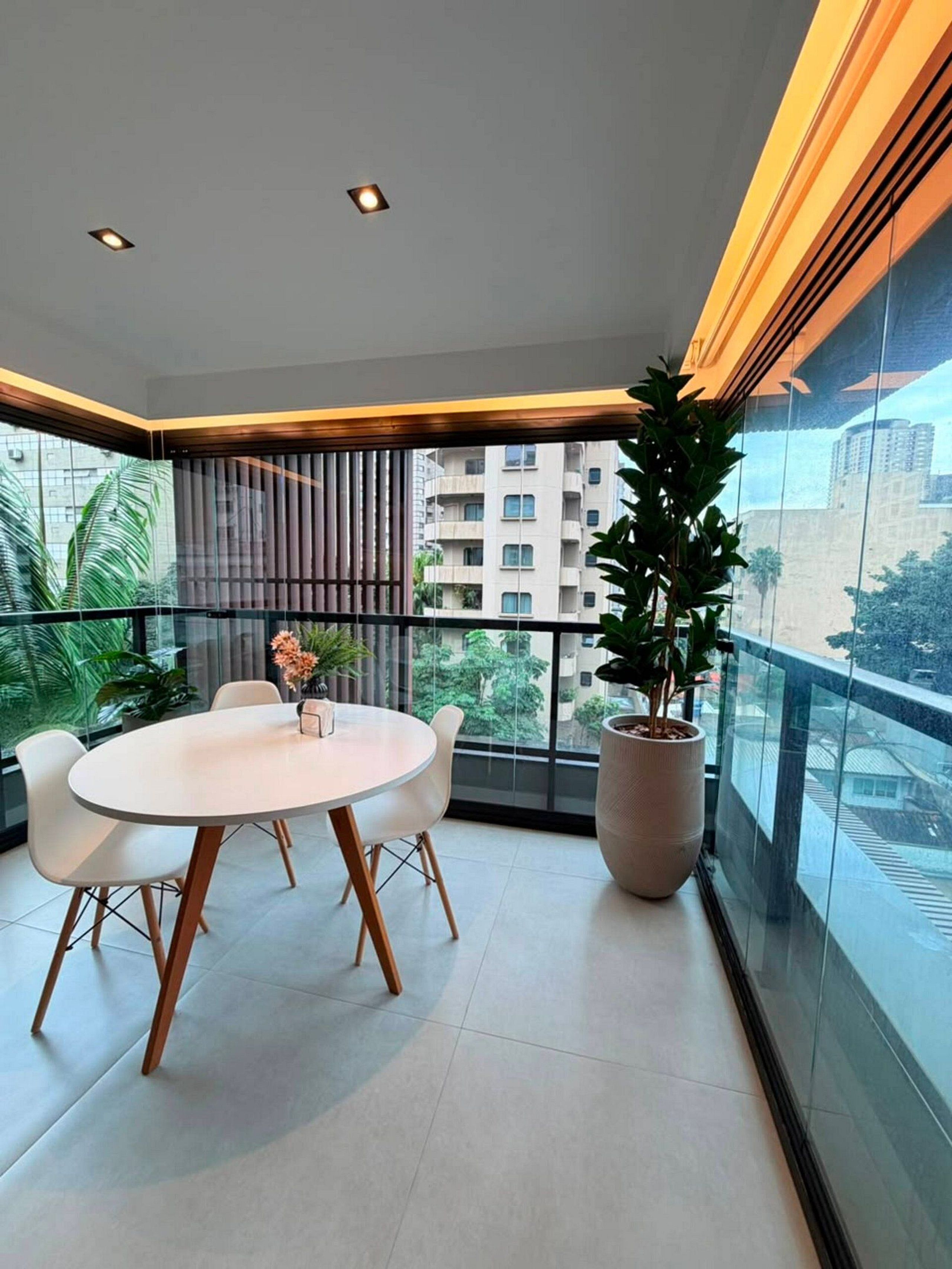A modern balcony with a dining table and chairs, a large potted plant, and a view of a cityscape.