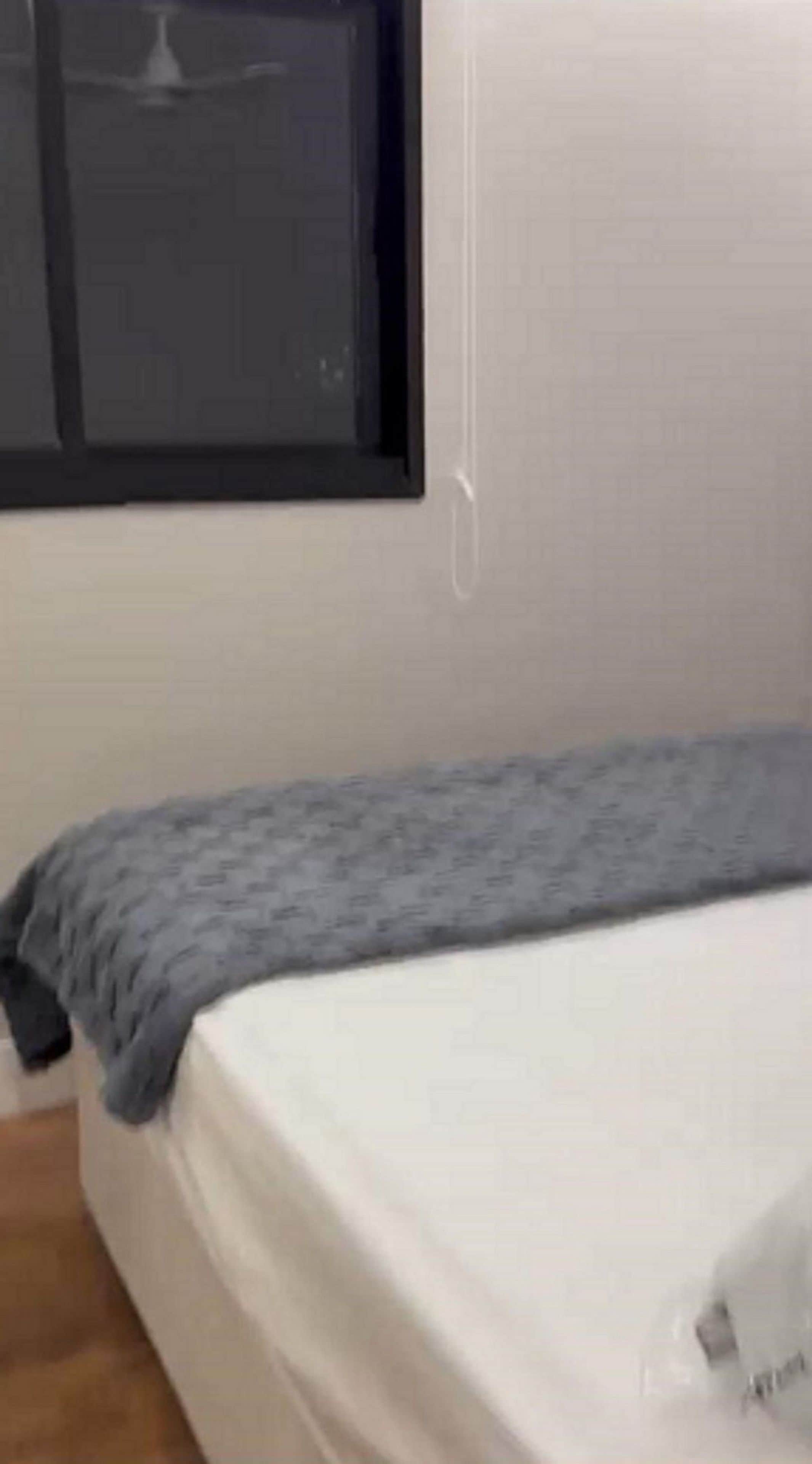 Un lit simple avec un matelas blanc et une couverture grise, une fenêtre avec un store fermé et un interrupteur fixé au mur.