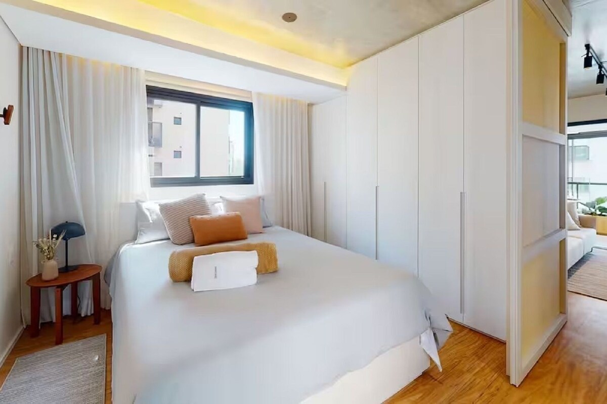 A modern bedroom with a large bed, a nightstand, a window, and a view of the city.