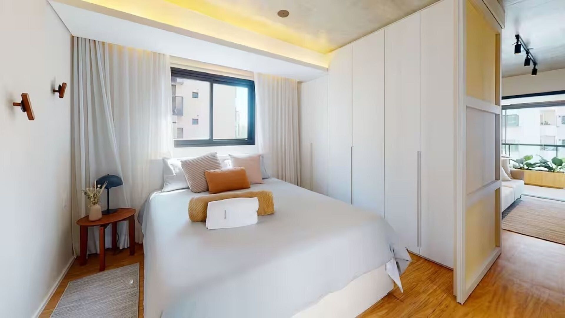 A modern bedroom with a large bed, a nightstand, a window, and a view of the city.