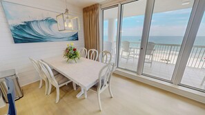 Uma área de refeições com mesa, cadeiras, um arranjo floral, uma pintura de uma onda e vista para a praia através de grandes janelas.