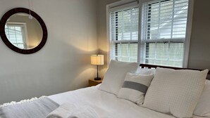 Ein ordentlich bezogenes Bett mit weißen Leinen, ein runder Spiegel an der Wand, eine Nachttischlampe und ein Fenster mit Jalousien.