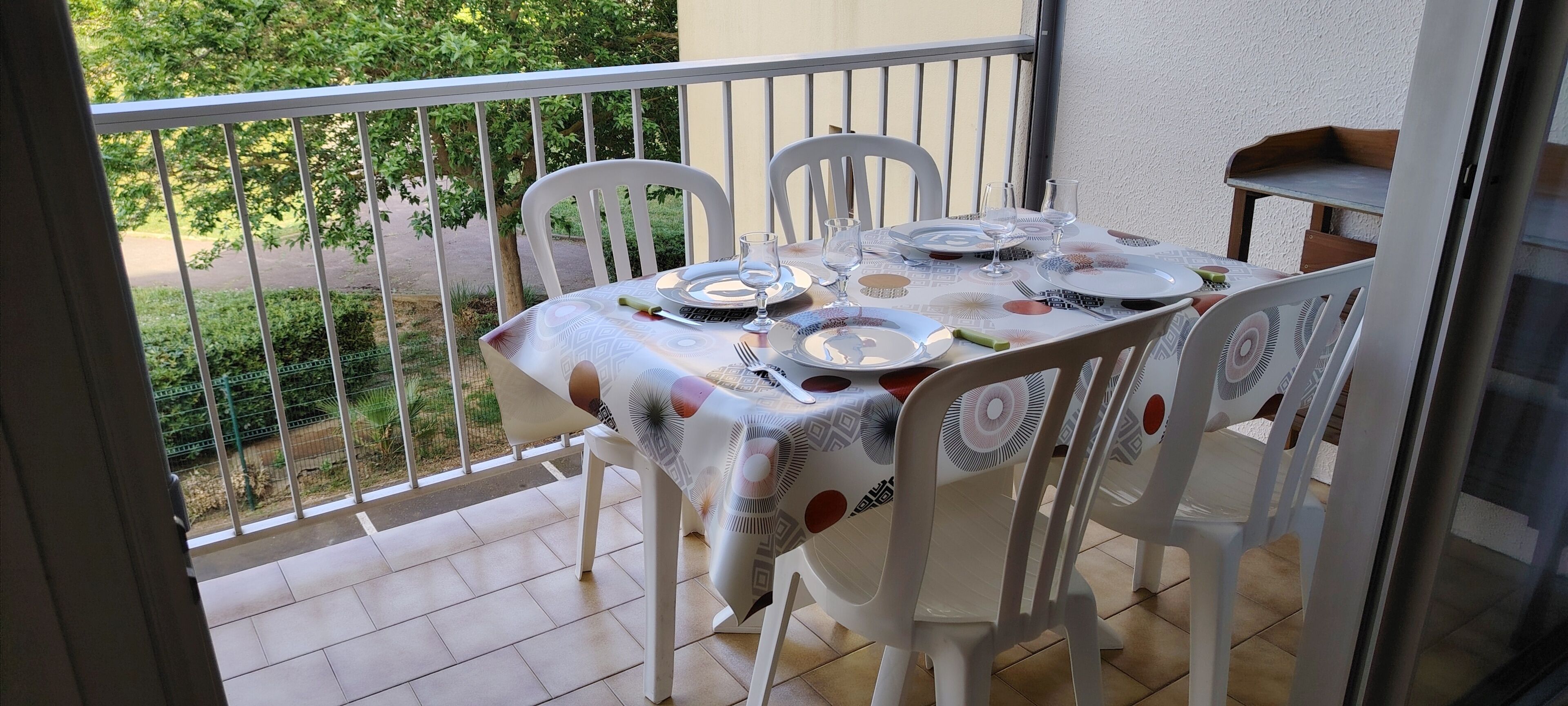A balcony with a table set for dining, white chairs, and a view of greenery.