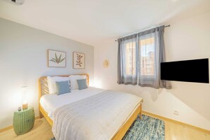Un dormitorio con cama, mesita de noche, televisor montado en la pared y ventana con cortinas.