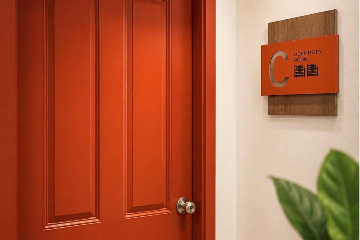Une porte rouge avec un trou de serrure, une pancarte indiquant une chambre de dortoir et un pot de fleurs sur la droite.