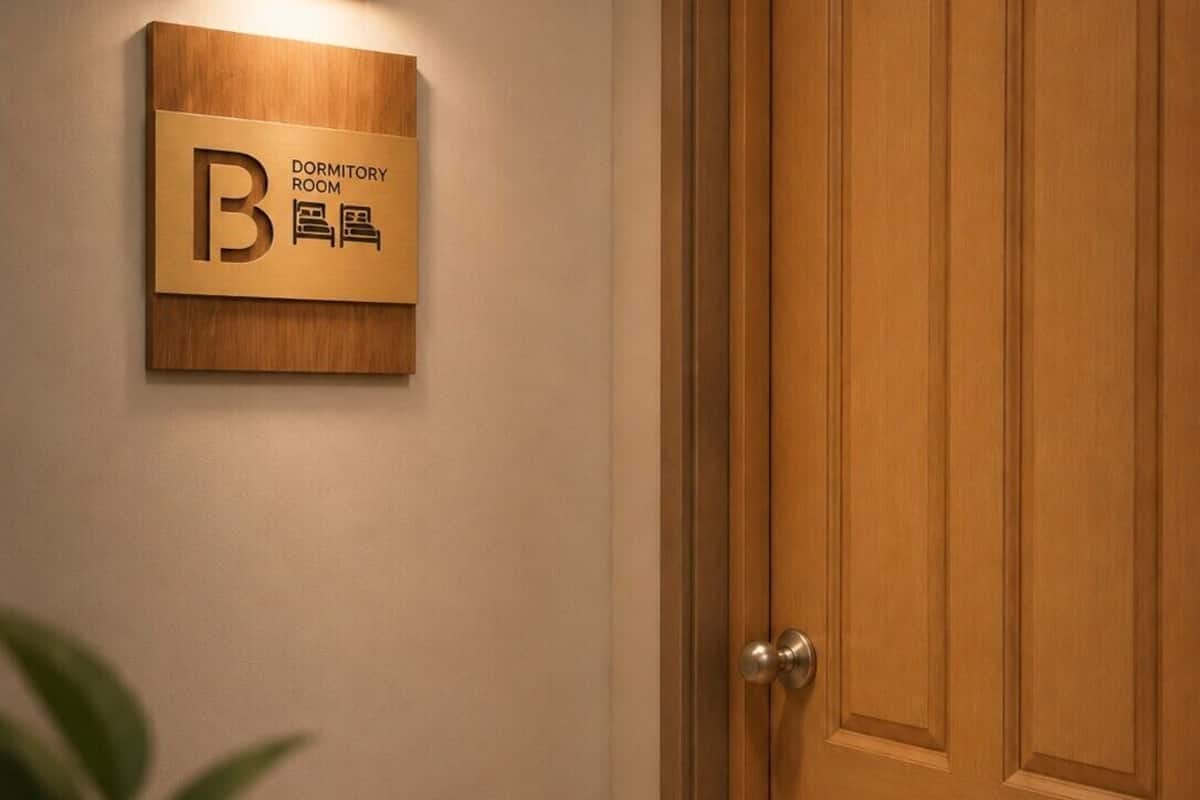 Une porte en bois avec une pancarte indiquant qu’il s’agit d’une chambre de dortoir, un luminaire fixé au mur et une plante au premier plan.