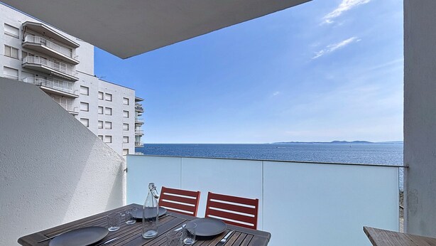 A balcony with a dining table set for two, overlooking a body of water and a distant landmass. - MEDA CHICA Roses / Roses - INTERimmo (Rosas / Roses)
