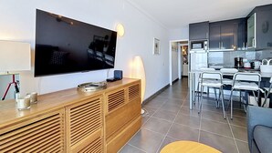 A modern living room with a flat-screen TV, wooden entertainment unit, and a dining area with a kitchen in the background. - MEDA CHICA Roses / Roses - INTERimmo (Rosas / Roses)