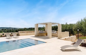 Ein Pool mit Liegestühlen und einer Pergola, umgeben von Steinmauern und Grünpflanzen.