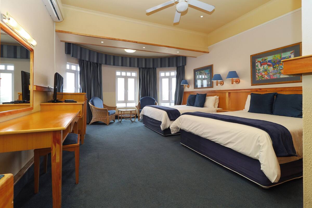 A hotel room with two beds, a desk, a chair, a television, and a ceiling fan.