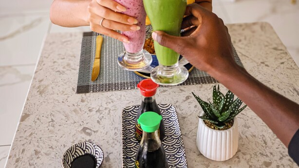 A person holding two smoothies at a table with a tray of condiments and a small potted plant.