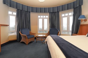 Quarto de hotel com cama, duas cadeiras, uma mesinha e amplas janelas com cortinas.
