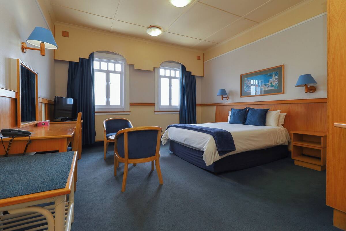 A hotel room with a bed, desk, chair, and a painting on the wall.