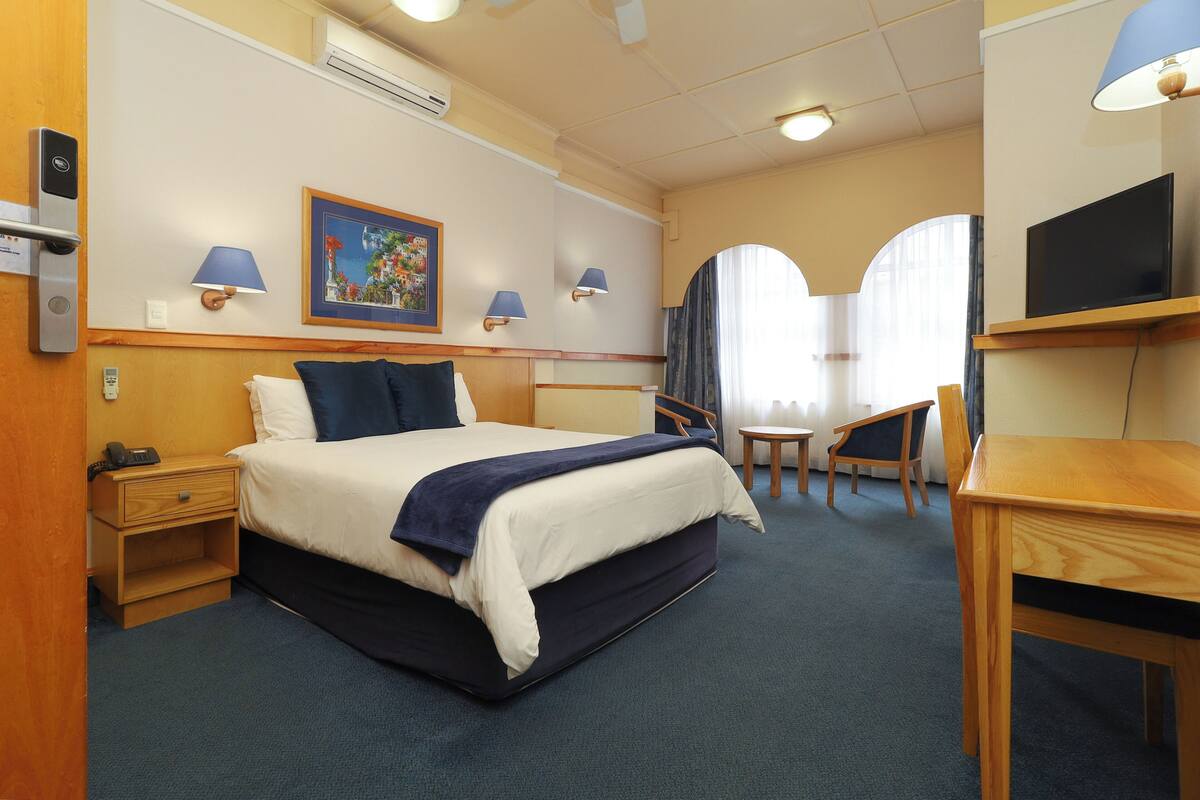 A hotel room with a bed, bedside tables, a desk, a chair, lamps, a TV, and a painting on the wall.