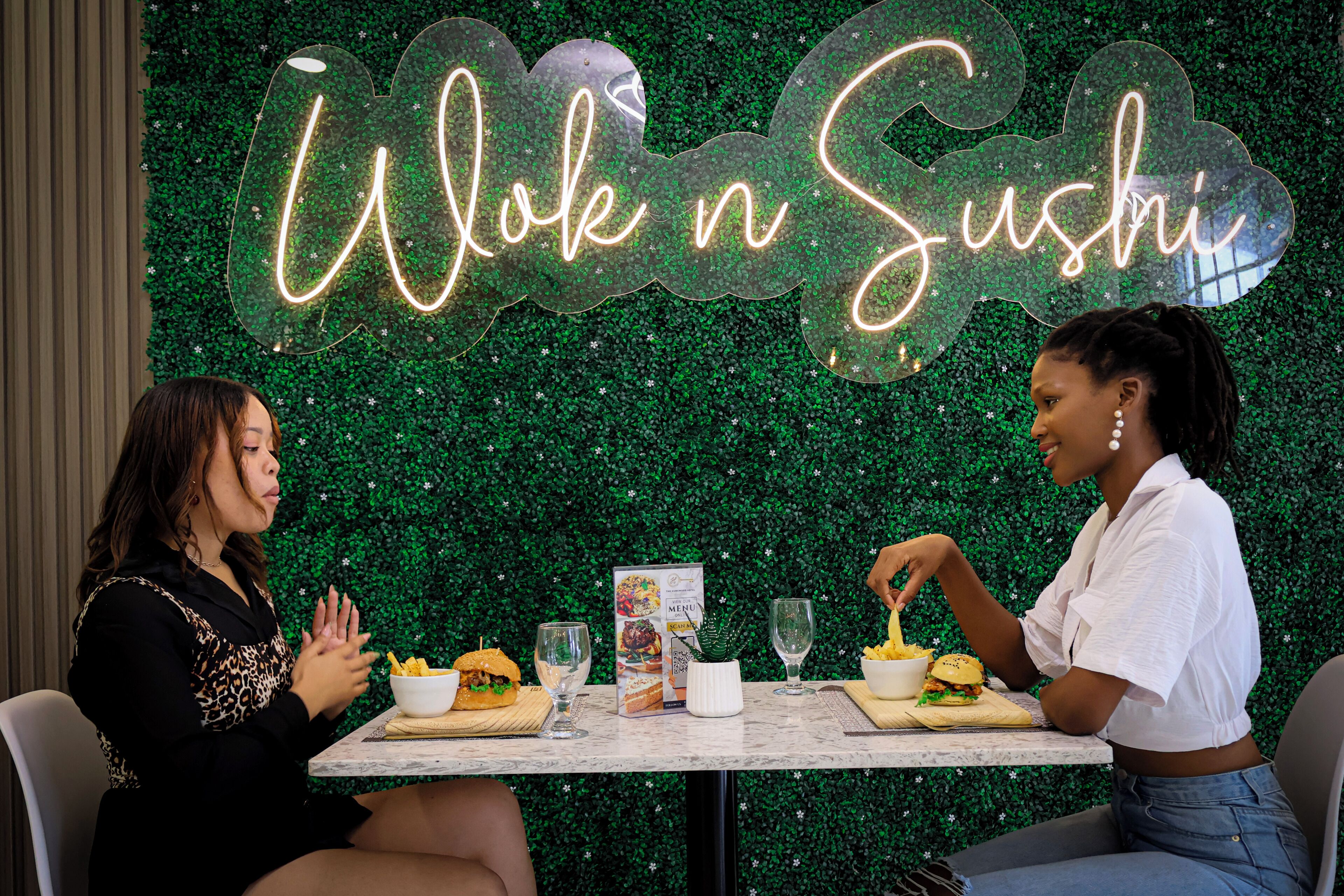 Two people seated at a table with food and drinks, a menu, and a neon sign reading 'Wok n Sushi' against a green leafy backdrop.
