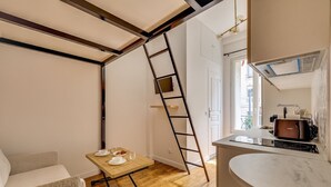 Een kamer met houten vloer, een bed, een tafel met stoel, een keukenhoek met aanrecht en apparatuur, en een ladder tegen de muur.