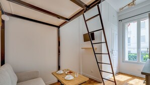 Een kamer met houten vloer, een witte bank, een houten tafel met dienblad, een ladder en een raam met uitzicht op gebouwen.