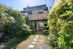 Un chemin de jardin menant à un bâtiment doté d’un balcon, entouré d’une végétation luxuriante et de plantes en pot.