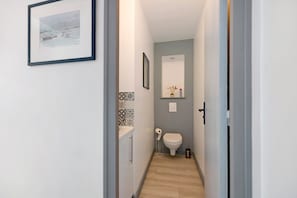 Un couloir étroit mène à une salle de bain équipée de toilettes, d’un distributeur de serviettes et d’un tableau encadré accroché au mur.