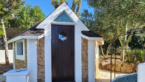 Um pequeno edifício de paredes de pedra com uma porta marrom escura, um pequeno alpendre e um muro de pedra.