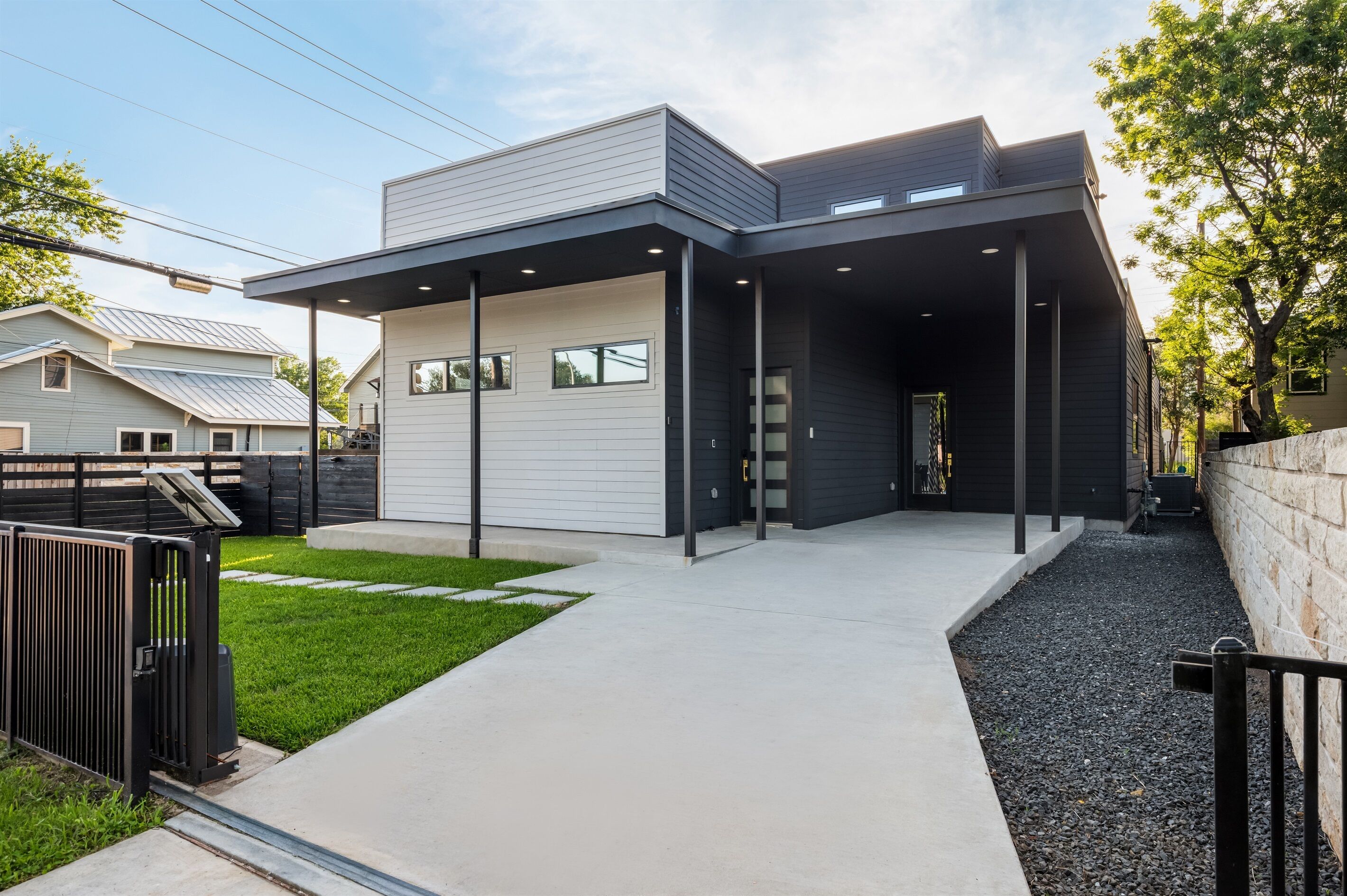 Une maison moderne à deux étages, dotée d’un patio couvert, d’une petite pelouse à l’avant et d’un mur en pierre sur le côté droit.