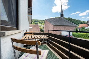 Ein Balkon mit einer hölzernen Bank und einem gepolsterten Stuhl, Blick auf einen Kirchturm und ein Backsteinhaus im Hintergrund.