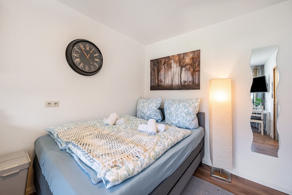 A neatly made bed with a patterned bedspread, a wall clock, a painting, a mirror, and a bedside lamp.