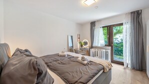 Un dormitorio con una cama grande, una mesita de noche con espejo, un radiador y una ventana con cortinas.