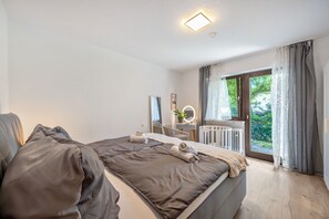 A bedroom with a large bed, a bedside table with a mirror, a radiator, and a window with curtains.