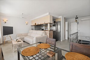 Ein modernes Wohnzimmer mit einer Couch, einem Glastisch mit gewebten Untersetzern, einer Küchenzeile mit Kühlschrank und einer Treppe mit Metallgeländern.