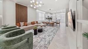 A modern living room with a sofa, coffee table, and chairs, a dining area with a table and chairs, and a kitchen in the background. - 5BR house sleeps 14 near Disney area (Clermont)