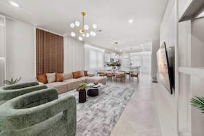 A modern living room with a sofa, coffee table, and chairs, a dining area with a table and chairs, and a kitchen in the background. - 5BR house sleeps 14 near Disney area (Clermont)