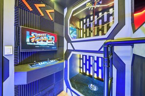 Ein Zimmer mit einem großen Bildschirm, der das Spiel 'Street Fighter: The Anniversary Edition' zeigt, einer Bedienkonsole mit Knöpfen und einem einzigartigen, geometrischen Wanddesign.