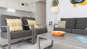Un salon moderne avec deux canapés gris, des coussins à motifs, une table basse en verre et, au fond, un espace cuisine.