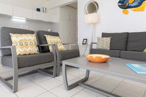 A modern living room with two grey sofas, patterned cushions, a glass coffee table, and a kitchen area in the background. - Belair Caribbean Sea with spectacular ocean views and house reef directly in front of the studio (Kralendijk)