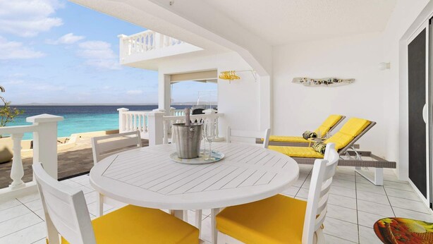 Un espace extérieur aménagé avec des meubles blancs, comprenant une table et des chaises, offrant une vue sur l’océan et doté d’un balcon avec des coussins jaunes.