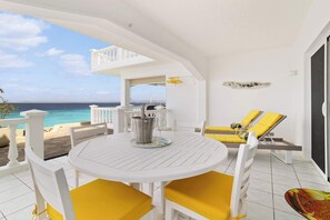 Un espace extérieur aménagé avec des meubles blancs, comprenant une table et des chaises, offrant une vue sur l’océan et doté d’un balcon avec des coussins jaunes.