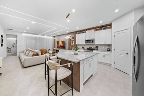 Una cocina moderna con armarios blancos, una isla central, taburetes de bar y un comedor con sofá y sillas.