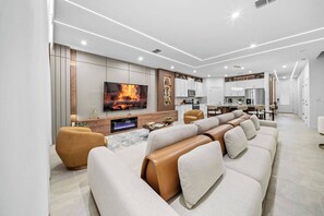A modern living room with a fireplace, sofa, TV, and kitchen area. - 8BR house with private pool in Solara Resort (Kissimmee)