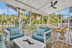 A covered outdoor seating area with white furniture, a ceiling fan, and a view of palm trees.