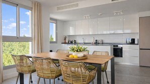Een moderne keuken met witte kasten, een houten eettafel, rieten stoelen en uitzicht op groen.