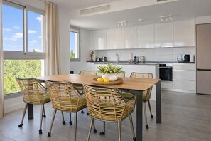A modern kitchen with white cabinets, a wooden dining table, wicker chairs, and a view of greenery outside. - Luxury Sea, Pool & Mountain View Apt near La Cala (Mijas Costa)
