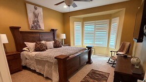 Un dormitorio con cama, mesitas de noche, ventilador de techo, ventana con persianas, alfombra y cómoda.