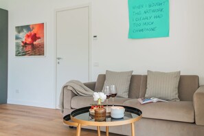 A living room with a beige sofa, a glass coffee table with a candle, wine glasses, and a framed artwork on the wall.