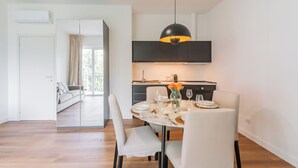 Un espace repas moderne avec une table ronde, des chaises blanches, un luminaire suspendu noir et une cuisine dotée d’un meuble noir et d’un téléviseur à écran plat.