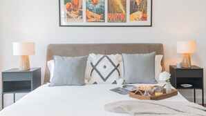 A neatly made bed with pillows, a tray with pastries and a glass of juice, and framed posters on the wall.