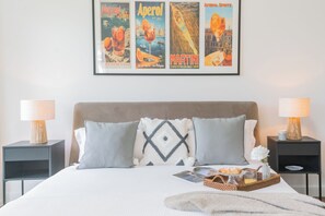 A neatly made bed with pillows, a tray with pastries and a glass of juice, and framed posters on the wall.